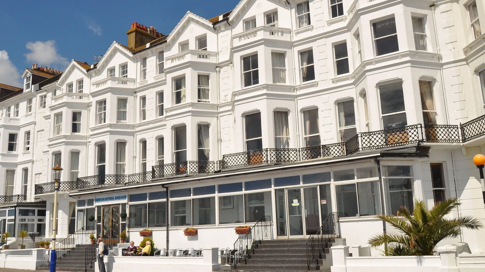 The white-rendered seafront facade of The Strand Hotel on Royal Parade, Eastbourne, with wrought-iron balconies and a glazed ground-floor sun lounge